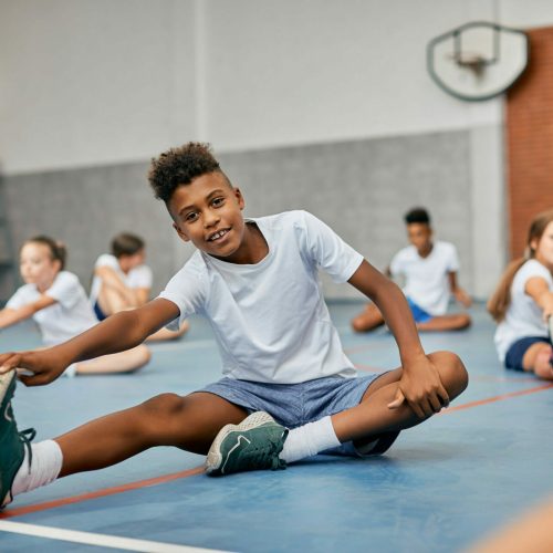 Happy African American schoolboy stretching during PE class at school gym.